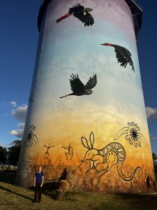 Ella stands in front of a water tower that has artwork featuring native Australian animals. 