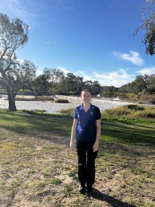 Ella stands and smiles to camera, on a grassy foreground with a scenic river in the background. 