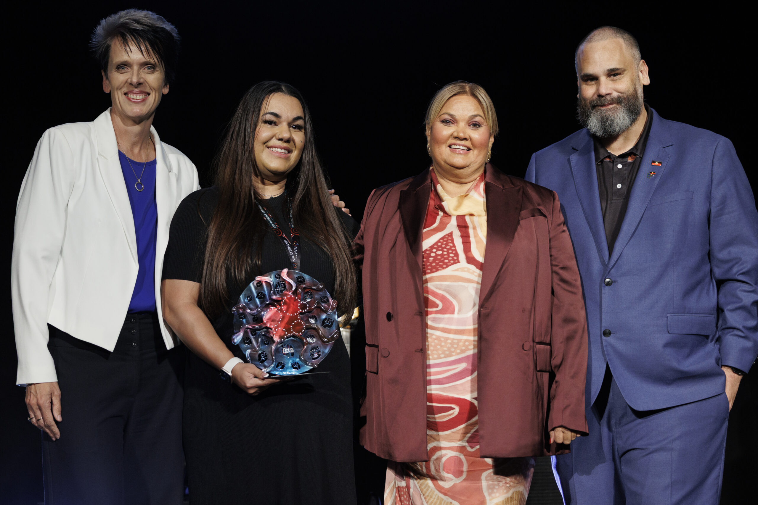4 people stand smiling at the camera. the person second from the left is holding a trophy.