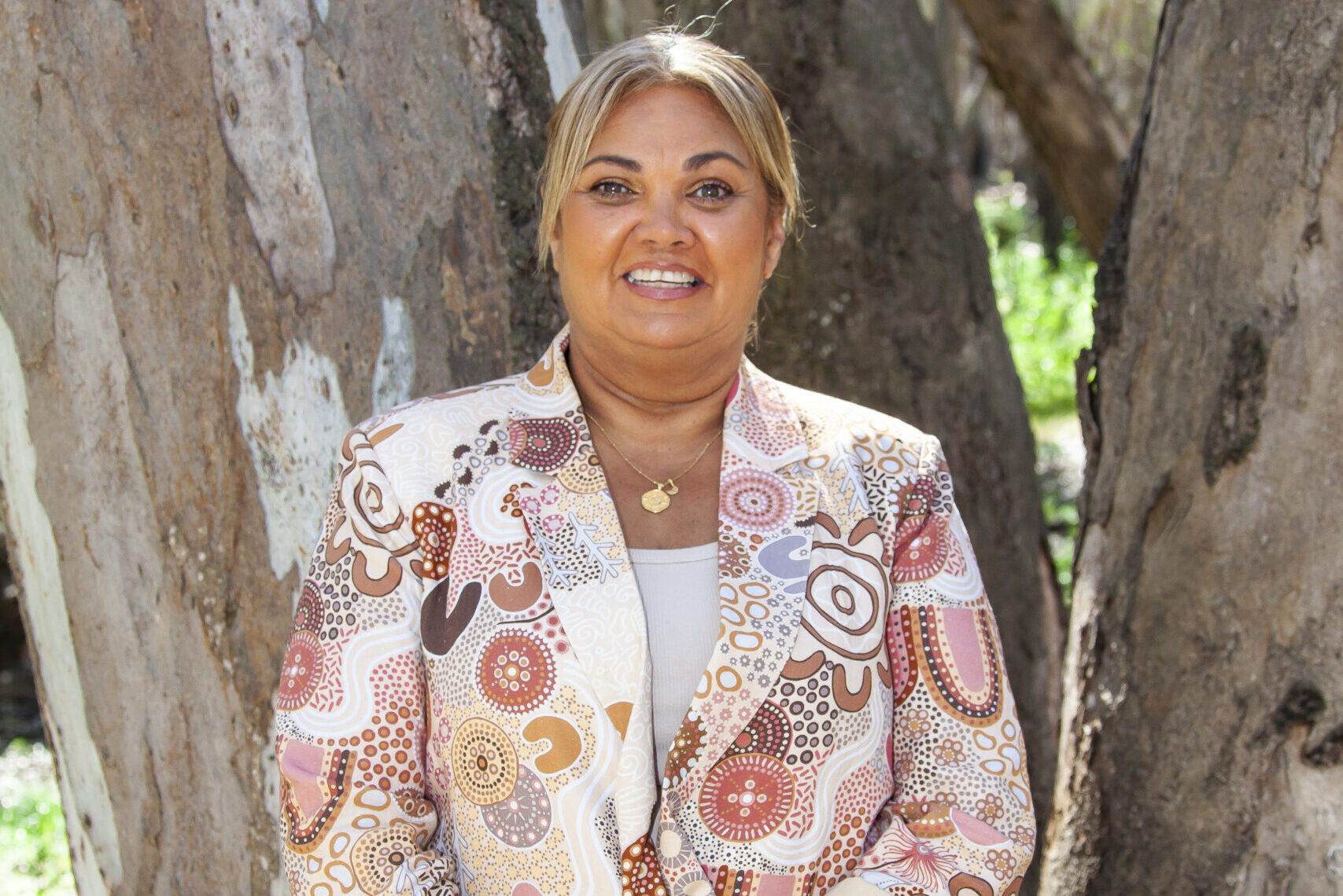 Karen stands in front of a tree, smiling at the camera and wearing a jacket with a first nations pattern on it.