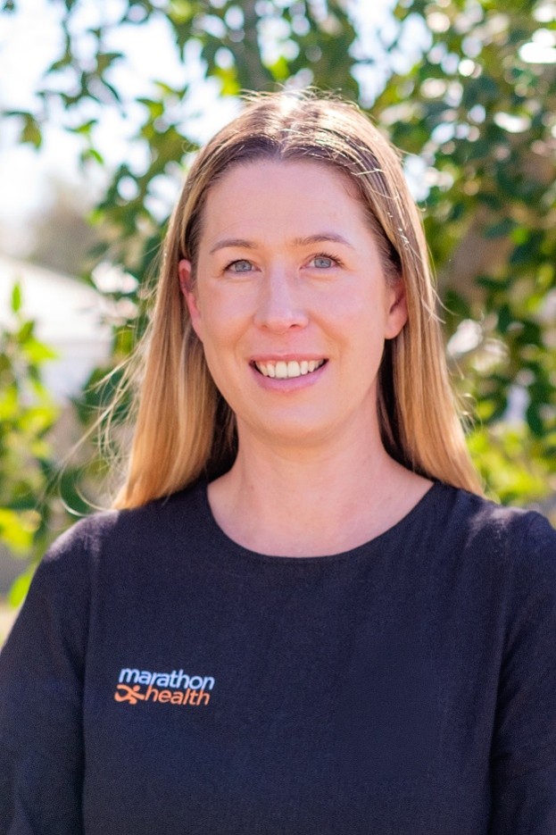 Kylie stands in front of leafy trees, wearing a Marathon Health shirt and smiling at the camera